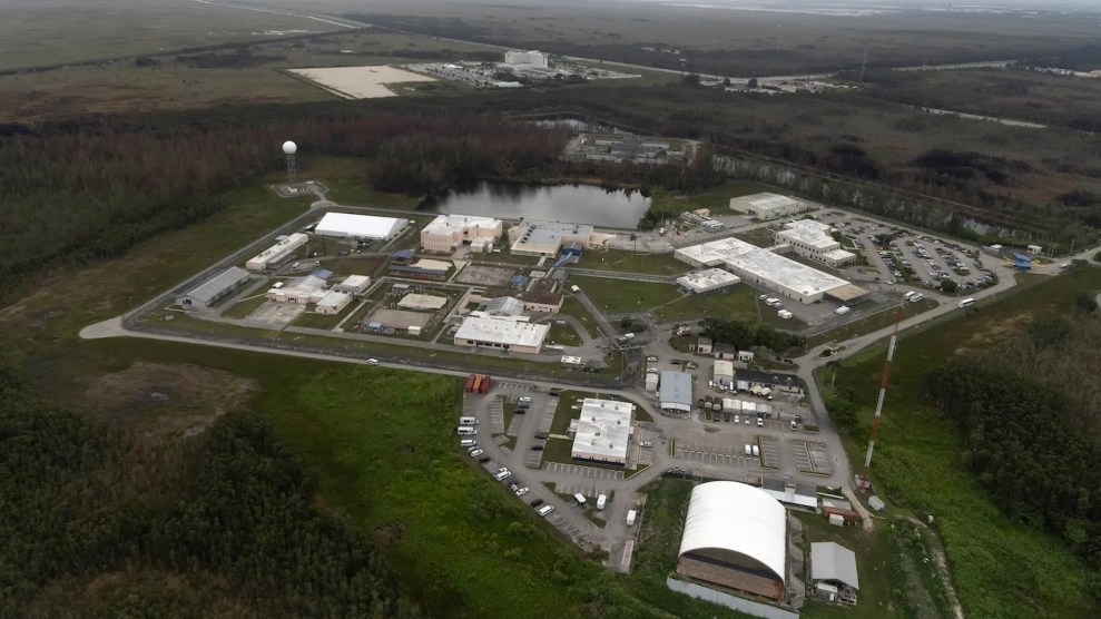 Aerial shot of white industrial buildings and parking lots surrounded by green grass and trees.